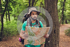 Male brunet bearded confused tourist got lost in the forest, holding map, looking far, trying to find the way. He has a backpack,