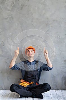 male builder in hard hat resting in lotus position and showing thumbs up at a place for text while sitting at work near