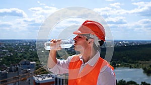 Male builder in helmet standing on the roof on the construction site and drinking water. Side view. Business, building