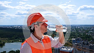 Male builder in helmet standing on the roof on the construction site and drinking water. Side view. Business, building