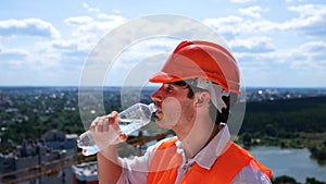 Male builder in helmet standing on the roof on the construction site and drinking water. Side view. Business, building