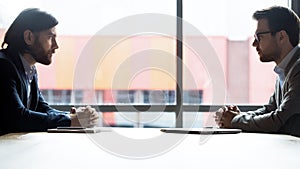 Male business opponents sit at desk looking at each other