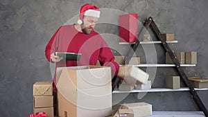 A male business owner is working packing an order to be sent to a client. wearing a santa claus hat and a red sweater