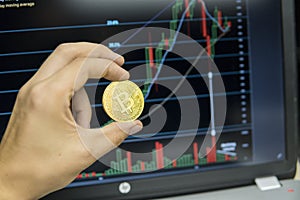 Male businessman hand holding bitcoin on a background of growth graph on a screen of laptop. Virtual money concept and