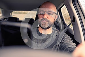 Male car driver in a car holding smart phone in his hand. The model is in his 40s, bald, with grey beard, dressed in grey shirt,