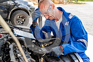 A male car mechanic wearing eye protection goggles in blue jumpsuit looking at the car engine thinking of the car problem and how