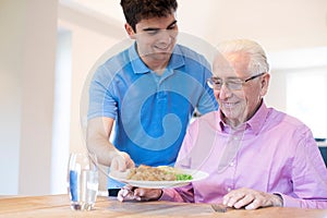 Male Care Assistant Serving Meal To Senior Male Seated At Table