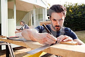 Male Carpenter With Female Apprentice Measuring Wood To Build Outdoor Summerhouse In Garden