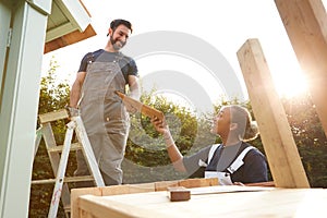 Male Carpenter With Female Apprentice Putting Roof On Outdoor Summerhouse In Garden