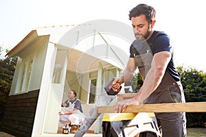 Male Carpenter With Female Apprentice Sawing Wood To Build Outdoor Summerhouse In Garden