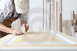 Male carpenter working the wood in carpentry workshop, putting paper masking tape on a wooden door, wearing overall and cap