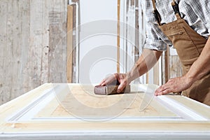 Male carpenter working the wood in carpentry workshop, sanding a wooden door with sandpaper, wearing overall