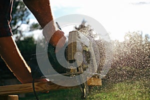 A male carpenter works with a circular saw on the lawn. Close-up view of a working tool. Contrast sunlight with glare scattered in