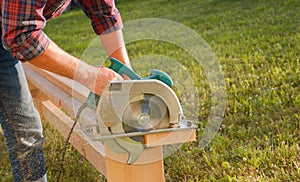 A male carpenter works with a circular saw on the lawn. Close-up view of a working tool. Contrast sunlight with glare scattered in