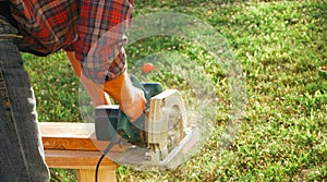A male carpenter works with a circular saw on the lawn. Close-up view of a working tool. Contrast sunlight with glare scattered in