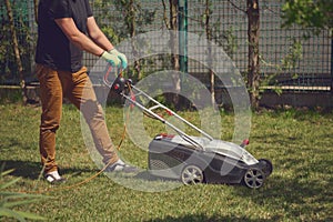 Male in casual outfit and gloves is cutting green grass with modern lawn mower on his backyard. Gardening care equipment