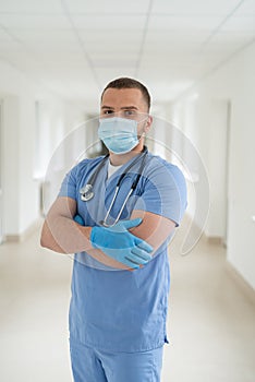 Male caucasian doctor wearing blue medical coat and face mask in modern clinic corridor