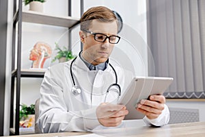 Male Caucasian doctor in white medical uniform filling patient medical history or anamnesis on tablet in office. Doctor consulting