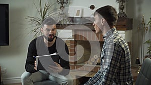 Male Caucasian psychologist in eyeglasses holding notes and talking to his sad patient. Young man undergoing