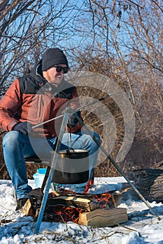 Male caucasian is sitting by campfire, pot of soot over bonfire hanging on tripod, winter outdoor cooking at campsite