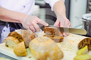 A male chef`s hand cutting whole grain bread with knife on the table with various kinds of freshly baked breads. Selective focus,