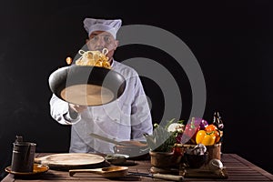 Male chef in white uniform holding a frying pan, sautÃ©ing spaghetti with fresh vegetables flying in the air