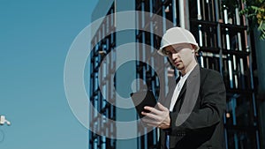 Male civil engineer contractor in safety helmet and jacket standing on construction site building and using mobile phone