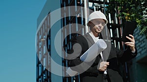 Male civil engineer contractor in safety helmet and jacket standing on construction site building and using mobile phone