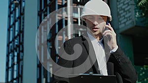 Male civil engineer contractor in safety helmet and jacket standing on construction site building and using mobile phone