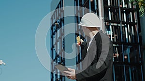 Male civil engineer contractor in safety helmet and jacket standing on construction site building and using mobile phone