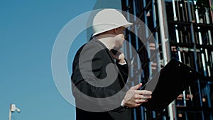 Male civil engineer contractor in safety helmet and jacket standing on construction site building and using mobile phone