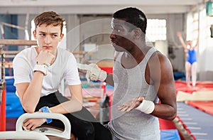 Male coach talking with upset teenage boy on gymnastic equipment at acrobatic center