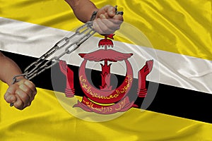 Male coarse hands chained in heavy iron chains against the background of the national flag of the state of Brunei, the concept of