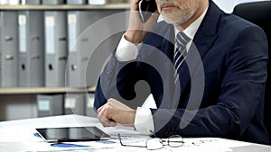 Male company director dialing, talking cell phone, tablet and glasses on table