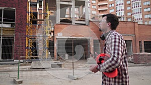 Male construction engineer walking through construction site and inspecting project, holding orange hard hat and use