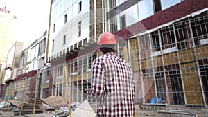Male construction engineer walking through construction site and inspecting project, holding orange hard hat and use