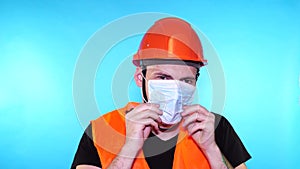 Male construction worker in overalls putting on medical mask on face on blue background.