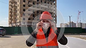 Male construction worker in overalls putting on medical mask on face on construction area.