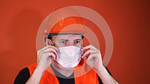 Male construction worker in overalls putting on medical mask on face on orange background.