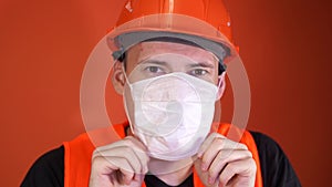 Male construction worker in overalls putting on medical mask on face on orange background.