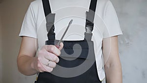 A male construction worker in overalls shows a flat screwdriver to the camera.