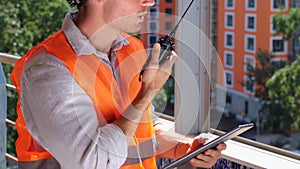 Male construction worker talking on walkie-talkie at construction site while holding digital tablet. Business, building