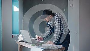 A male contractor builder at a construction site, working with a project on a laptop. An engineer is using blueprints to