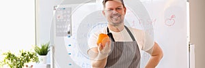 Male cook teacher in apron holds an orange and fresh vegetables and fruits on table