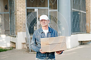 Male courier in the blue costume and a cap taking out mail carton boxes from the white van on the sunny day in the street. Outdoor photo