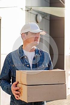 Male courier in the blue costume and a cap taking out mail carton boxes from the white van on the sunny day in the street. Outdoor photo