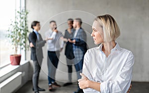 Male Coworkers Whispering Behind Back Of Unhappy Businesswoman In Office