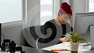 A male creative graphic designer is reading a book while sitting in front of computer.