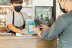 Male customer with protective mask paying bill by cell phone in cafe.