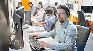 Male customer service representative with headset working in call center office with colleagues in row.